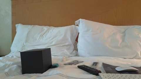Video of a neatly made bed with white pillows and a wooden headboard, featuring a black box, remote, glasses, and a white keyboard on the sheets.