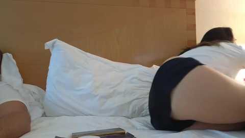 Video of a woman lying on a bed, wearing a white shirt and black shorts, head resting on a pillow, with a wooden headboard and white sheets in the background.
