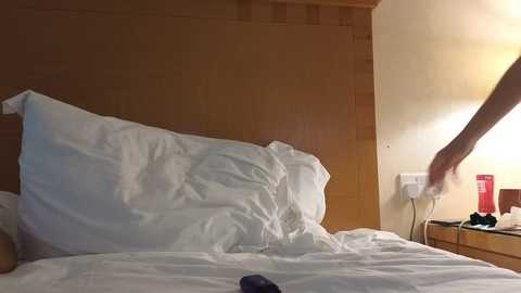 Video of a dimly-lit hotel room with a large, unmade bed featuring white pillows and sheets. A person\u2019s arm extends into the frame from the right side, reaching for something on a wooden nightstand. The background shows a beige wall and a bedside lamp.