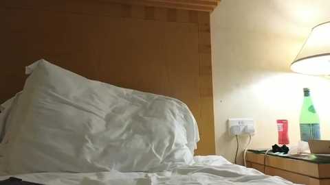 A video of a cozy bedroom corner, featuring a wooden headboard, a rumpled white duvet, a bedside lamp with a green bottle, and a red can, casting a warm, inviting glow.