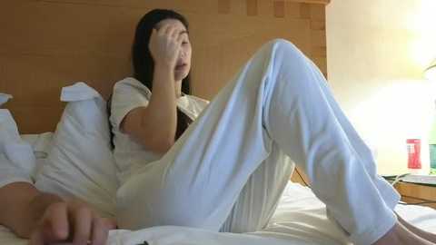 Video of an Asian woman with long black hair, wearing white pajamas, sitting on a bed, covering her face with her hands, in a warmly lit, wooden-paneled bedroom.