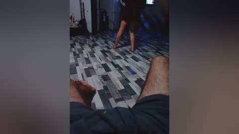 Video of a dimly lit room with a man in a dark t-shirt and shorts, standing on a gray and black patterned carpet.