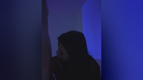 Video of a young woman with shoulder-length dark hair, wearing a red top, crouched in a dimly lit, narrow hallway. The background is blue, suggesting a moody, suspenseful atmosphere.