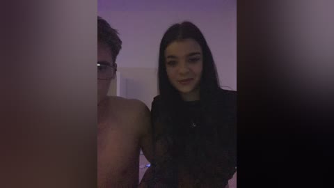 Video of a shirtless man with short curly hair, standing in front of a woman with long dark hair, wearing a black lace top, in a dimly lit room.
