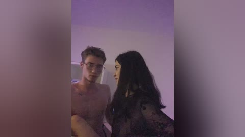 A video shows a young man with short blonde hair and glasses, wearing a white shirt, and a woman with long black hair, dressed in a black lace top, in a dimly lit room.