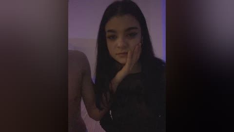 A dimly lit video of a young woman with long black hair, wearing a black top, touching her face, standing in front of a blurred background.
