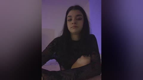 Video of a young woman with long dark hair, wearing a sheer black lace top, partially exposing her breast. She has a neutral expression. The background is dimly lit with purple and blue hues.