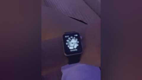 A video shows a darkened room with a rectangular, dimly lit Apple Watch on a white surface. The watch face displays a blurred, white clock-like image.