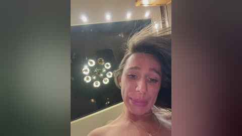 A video of a smiling woman with long brown hair, wearing a gold necklace, taken indoors with warm lighting, showing her in a mirror reflection.