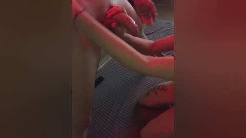 Video of a woman with fair skin and red hair, partially naked, holding a red dildo. She is wearing a grey knitted sweater and has a tattoo on her right forearm. The scene is dimly lit with red and blue lighting.