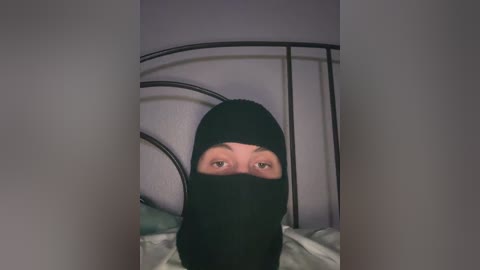 Video of a person in a black hooded sweatshirt, with eyes visible through the hood's eye openings, standing in a dimly lit room with a black metal bed frame.