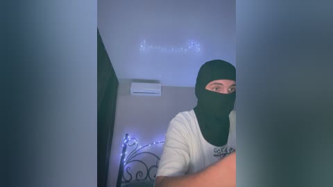 Video of a man in a black balaclava, white T-shirt, and black gloves, standing in a dimly lit room with an air conditioner and decorative lights on the wall.