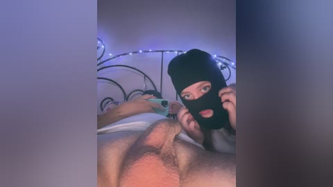 Video of a light-skinned man with a black beanie and black face mask, lying on a bed, exposing his erect penis, with a woman with long dark hair and a green phone in the background.