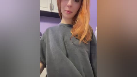 Video of a young woman with light skin and shoulder-length, dyed orange hair, wearing a gray T-shirt, standing in a room with white cabinets and purple walls.
