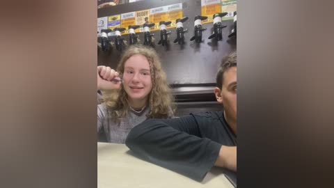 Media: Video of a young girl with curly blonde hair, smiling and giving a thumbs-up, seated next to a boy in a dark shirt at a restaurant with a beer tap display in the background.