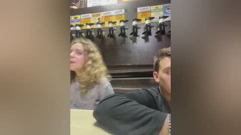 Media: Video of a man and woman with curly hair, blurred, sitting in a restaurant booth with a wall of black hats, yellow signs, and a dark background.