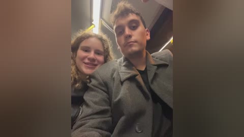 Media: Video of a young couple sitting close together on a train. The woman, with curly blonde hair, wears a grey coat, smiling. The man, with short brown hair, wears a grey coat, black shirt, and looks at the camera.