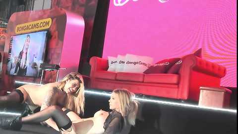 Media: Video of two nude women in lingerie, one performing oral sex on the other, in a brightly lit, modern living room with red sofa and abstract pink wall art.
