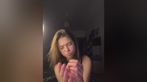 Video of a young woman with light skin and shoulder-length blonde hair, wearing a black top, squinting and cupping her hands in front of her face in a dimly lit room.