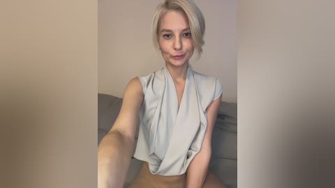 Video of a fair-skinned, blonde woman with a short bob haircut, wearing a sleeveless, light-gray top. She sits on a gray couch against a beige wall, smiling slightly.