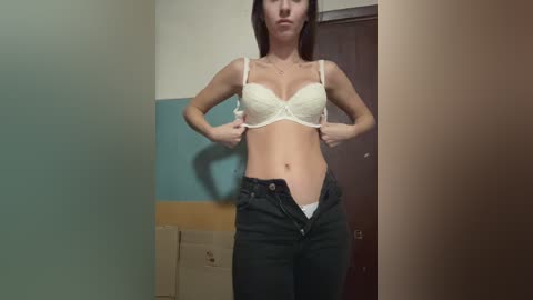 Media: Video of a light-skinned woman with brown hair, wearing a white lace bra and black jeans, partially unzipped, standing in a dimly lit room with beige walls and a wooden door.