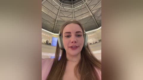 Media: Video of a young, fair-skinned woman with long, straight, light brown hair wearing light green headphones, standing in a modern, circular, glass-enclosed room with a high, metallic ceiling.