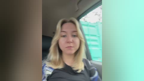 Video of a blonde woman with a neutral expression, wearing a dark shirt, seated in a car. Background features a turquoise-green container and blurred foliage.