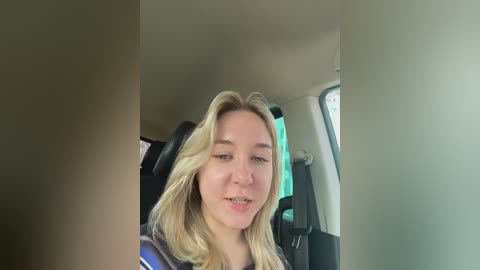Video of a young, fair-skinned woman with long, wavy blonde hair, sitting in a car, wearing a blue shirt. She smiles slightly. The car's interior features dark leather seats and a greenish tinted window.