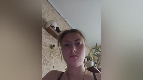 Video of a young woman with fair skin and blonde hair, wearing a black top, standing against a beige textured wall. A small potted plant and a shelf with a lamp are in the background.