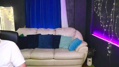 Video of a cozy living room with a white leather sofa adorned with blue and turquoise pillows. The background features dark curtains and glowing fairy lights, creating a warm, intimate atmosphere.