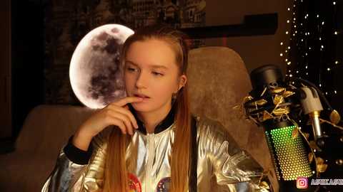 Video of a young woman with long blonde hair, wearing a metallic jacket, seated indoors, gazing pensively at a large full moon poster, surrounded by a cozy, dimly lit room with fairy lights.