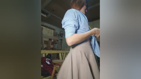 A video of a woman in a blue shirt and beige skirt, adjusting her skirt, standing in a cluttered room with a wooden chair, a red bag, and a bookshelf in the background.