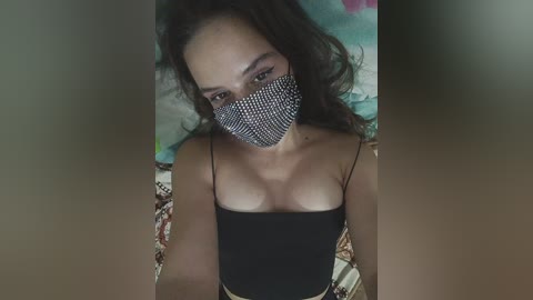 A video of a young woman with long, dark hair, wearing a black, spaghetti-strap top and a face mask with a black and white diamond pattern, lying on a colorful bedspread.