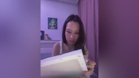 Media: A video of a young Asian woman with long black hair, wearing a white tank top, sitting on a bed. She's holding a white box, with a soft purple light illuminating the room.