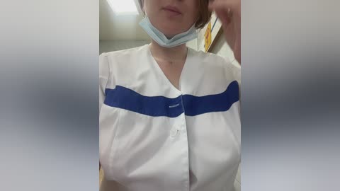 Video of a woman wearing a white surgical gown with a blue band, a light blue face mask, and a stethoscope around her neck. She stands indoors, partially blurred.