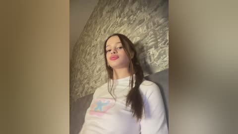 Video of a young woman with fair skin and long brown hair, wearing a white t-shirt with a blue star design, against a textured gray wall.