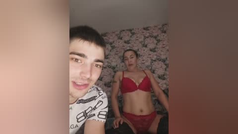 Video of a young man with short brown hair, wearing a white T-shirt, and a woman with blonde hair, wearing a red lace bra and panties, in a room with floral wallpaper.