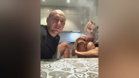 Media: Video of a bald, muscular man and a blonde woman with large breasts, sitting on a tiled kitchen counter, wearing black shirts, surrounded by steam.