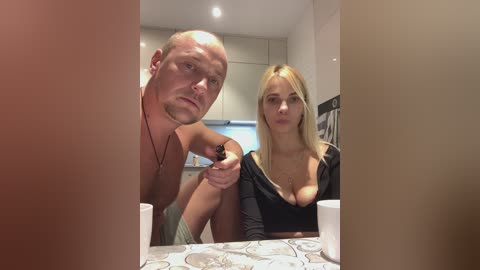 Media: Video of a blonde woman with large breasts in a low-cut black top, seated beside a bald, muscular man in a kitchen with beige walls and white cabinets.