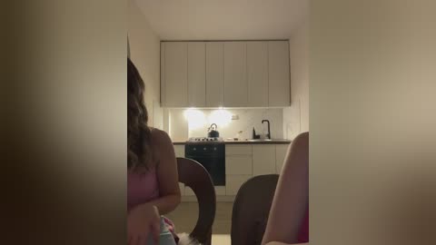 Media: A video of a dimly lit, minimalist kitchen with white cabinets, a black stove, and a sink, viewed from an angle, with a woman's back visible in a pink tank top.