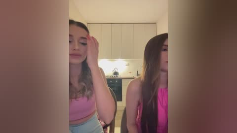 Media: Video of two young women with fair skin, one with long brown hair in a pink top, the other with long blonde hair in a pink top, standing in a modern kitchen with white cabinets and a countertop.