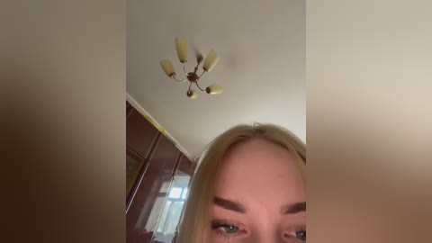 A video of a blonde woman with light skin and arched eyebrows, wearing makeup, standing indoors. The background shows a beige ceiling with a five-light chandelier and a partially visible, dark wooden wardrobe.