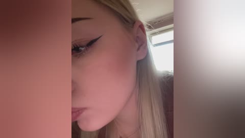 Close-up video of a young woman with long blonde hair, wearing heavy black eyeliner and light makeup, gazing to the right. Background shows a window with blinds.