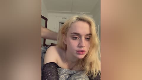 Video of a young, pale-skinned woman with shoulder-length blonde hair, wearing a black mesh top, looking intently at a mirror in a dimly lit bedroom.