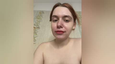 Video of a topless young Caucasian woman with fair skin, brown hair, and light makeup, standing in a bathroom with beige walls and a floral shower curtain.