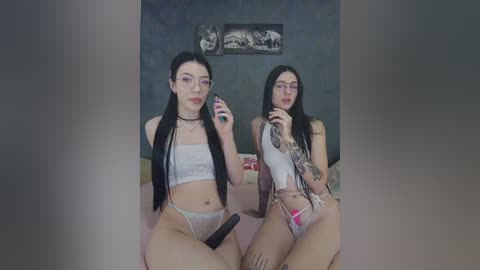 Video of two young women with pale skin, one wearing a choker and glasses, the other with tattoos, sitting on a bed, both holding dildos.