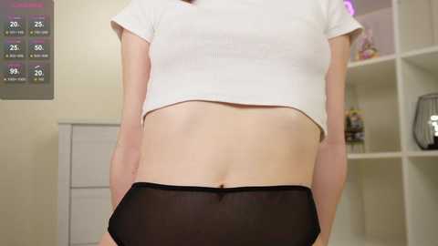 Video of a slim, light-skinned person wearing a white ribbed crop top and sheer black panties, standing in a well-lit room with a white shelf and a temperature display.