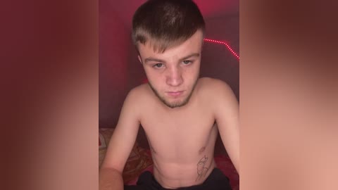 Video of a young, fair-skinned, bearded man with short brown hair, shirtless, sitting on a bed with a red light strip on the wall.