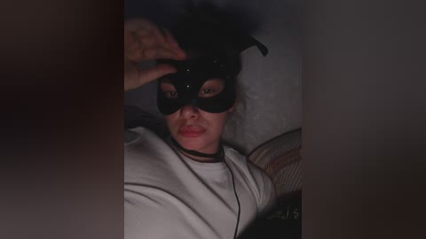 Media: Video of a woman wearing a black cat mask, lying on a bed, with a dimly lit room in the background.