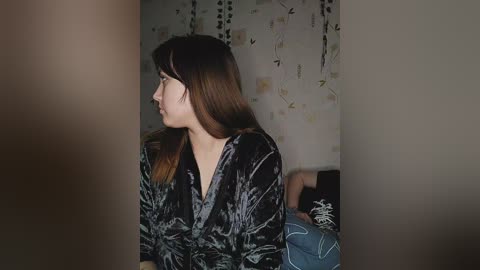 Media: Video of a young woman with light brown hair, wearing a black velvet top, sitting on a bed with a patterned duvet, against a floral wallpaper background.